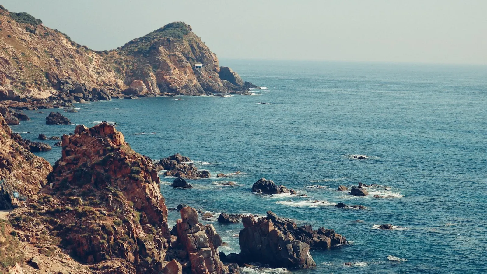 Bờ biển đá Quy Nhơn với những mỏm đá nâu nhô ra mặt nước xanh, xa xa là mũi núi phủ cây