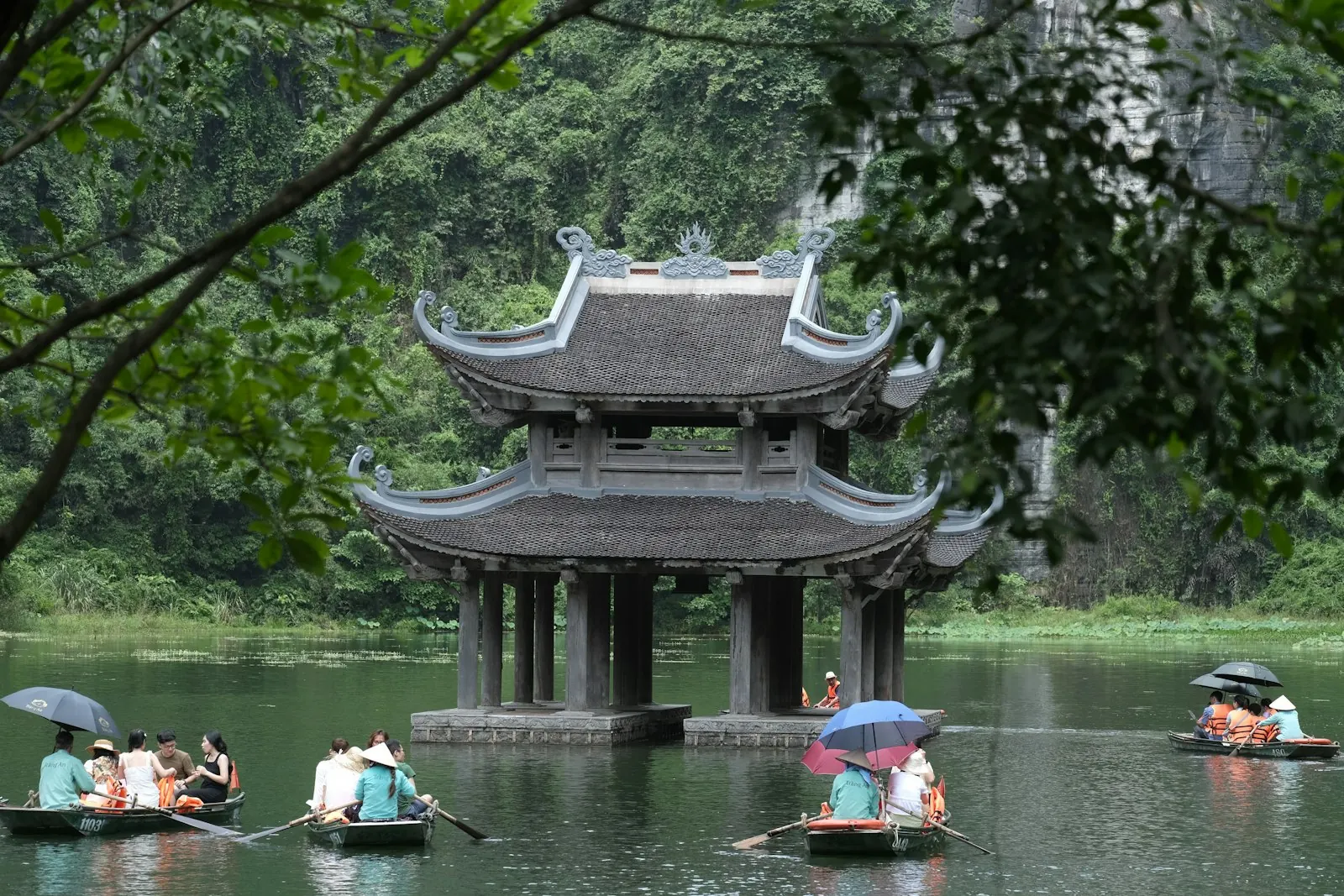 Ngôi đền đá mái cong dựng giữa mặt nước ở Tràng An, vài chiếc đò chèo tay chở khách đi ngang phía trước