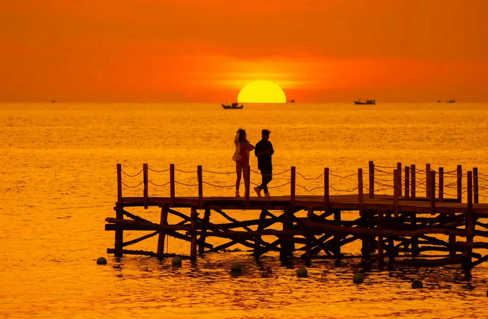 Hai người đứng trên cầu gỗ nhô ra biển ở Hòn Thơm lúc hoàng hôn, mặt trời đỏ lớn sát đường chân trời